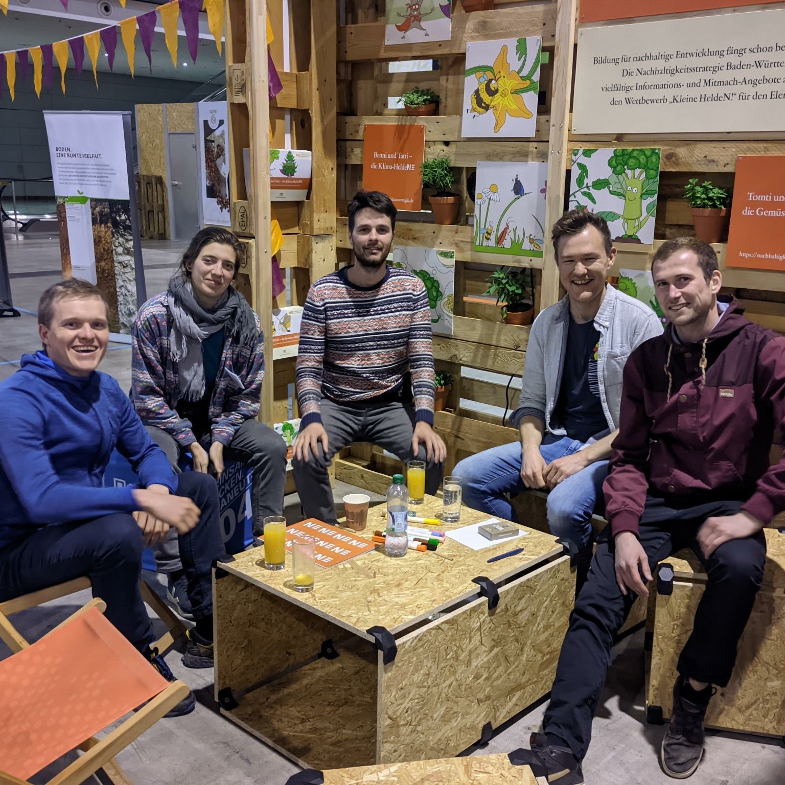A group of five people sitting around a table in a cozy, creative indoor setting, smiling and enjoying drinks.