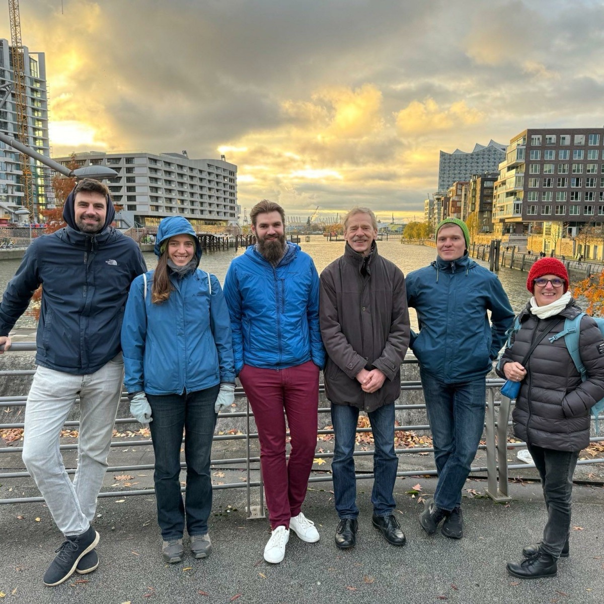 Group of six people smiling in front of a scenic urban waterfront at sunset, wearing winter clothing, conveying friendship and warmth.