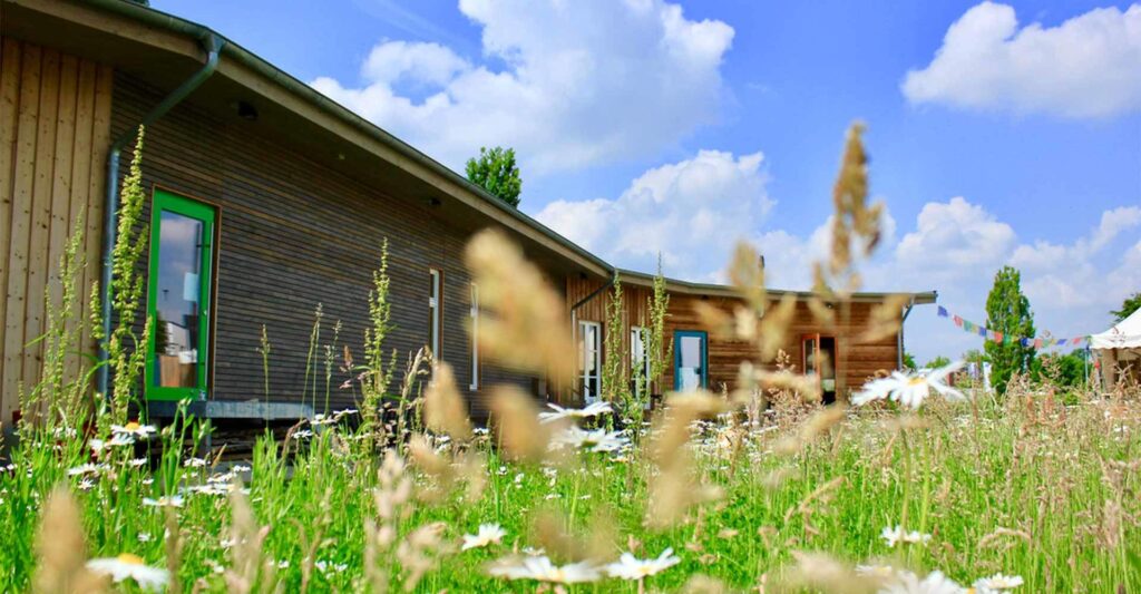 Das Kulturquartier Münster - ein einstöckiges Holzgebäude mit grünen Fensterrahmen, im Vordergrund Blumen und Gräser, im Hintergrund eine Girlande.