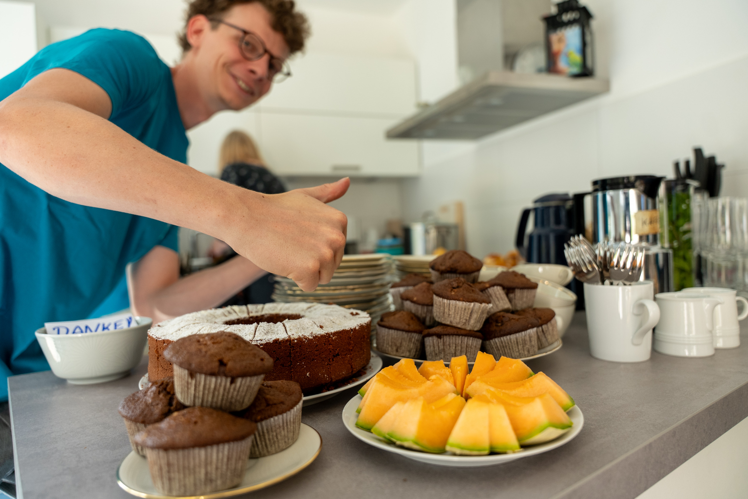 Auf einem Tisch stehen Kuchen, Muffins und fertig zugeschnittene Melonenstücke. Im Hintergrund ist ein Mitglied der Regionalgruppe zu sehen, das einen Daumen nach oben zeigt und fröhlich dazu lächelt.