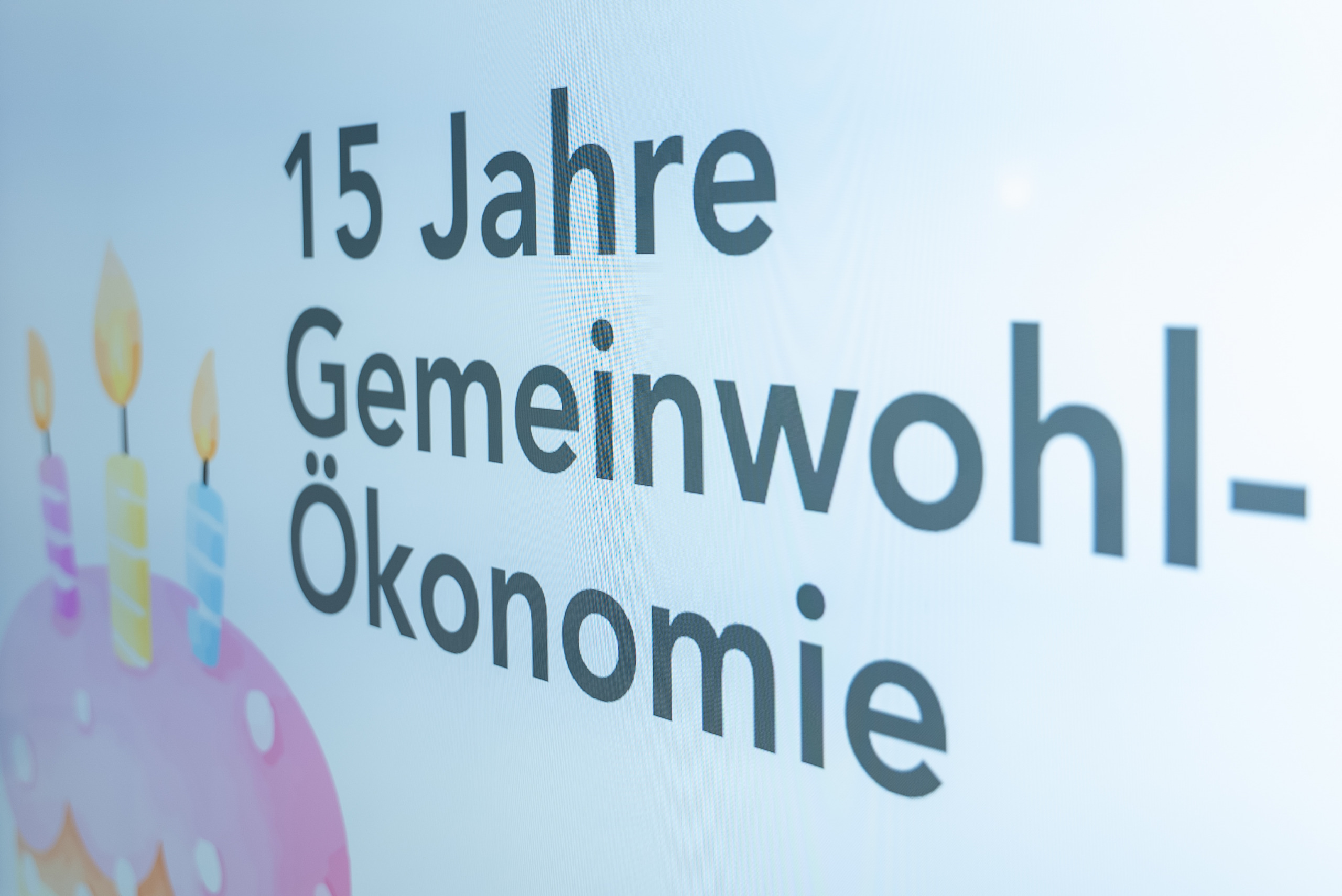Auf einer Leinwand ist neben einer stilisierten Geburtstagstorte folgender Text zu lesen: 15 Jahre Gemeinwohl-Ökonomie.