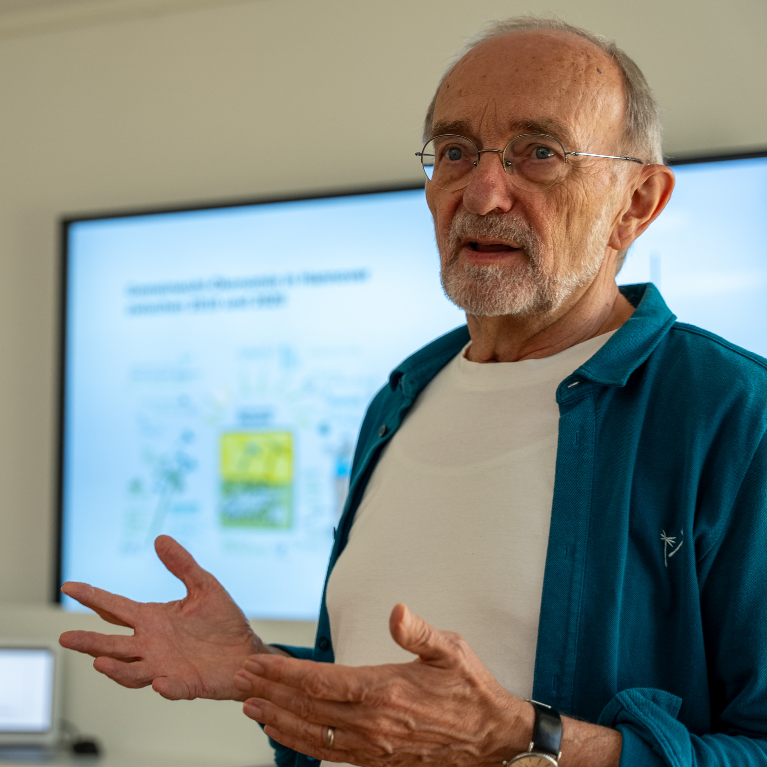 Günther Baumert steht in seinem Vortrag vor einer Leinwand, auf der eine PowerPoint-Präsentation gezeigt wird.
